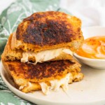 A halved, stacked grilled cheese sandwich on a plate with a bowl of dip.