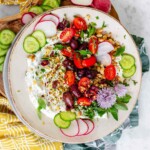 White dip on a plate topped with nuts, spices, tomatoes, black olives, sliced cucumber and radish, oregano leaves, and chive blossoms.