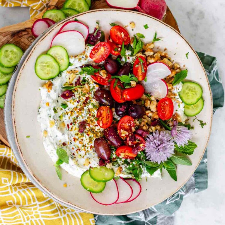 White dip on a plate topped with nuts, spices, tomatoes, black olives, sliced cucumber and radish, oregano leaves, and chive blossoms.