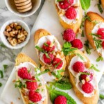 Toasted baguette slices topped with white spread, raspberries, chopped mint, walnuts, and honey.