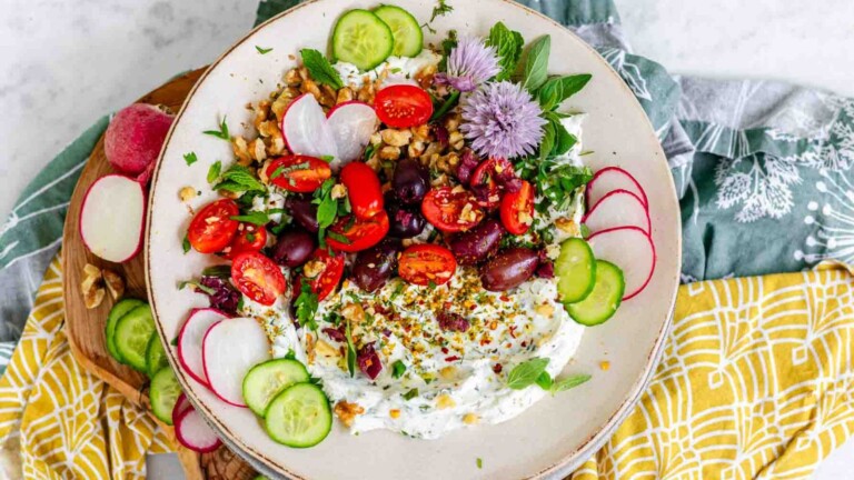 White dip on a plate topped with nuts, spices, tomatoes, black olives, sliced cucumber and radish, oregano leaves, and chive blossoms.