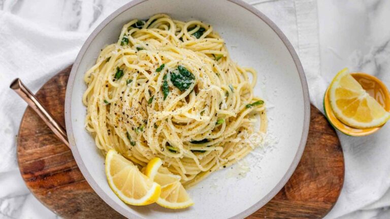 Pasta in a bowl with lemon wedges, topped with greens and cheese.