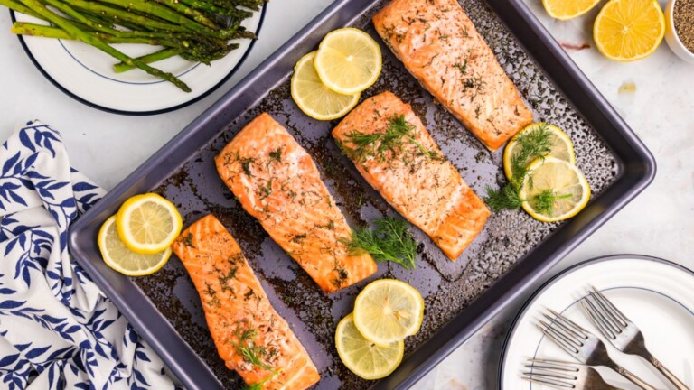 A sheet pan with four salmon fillets surrounded by lemon slices and dill.