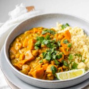 Curry with chunks of sweet potato in a bowl with couscous, a lime wedge, and cilantro.