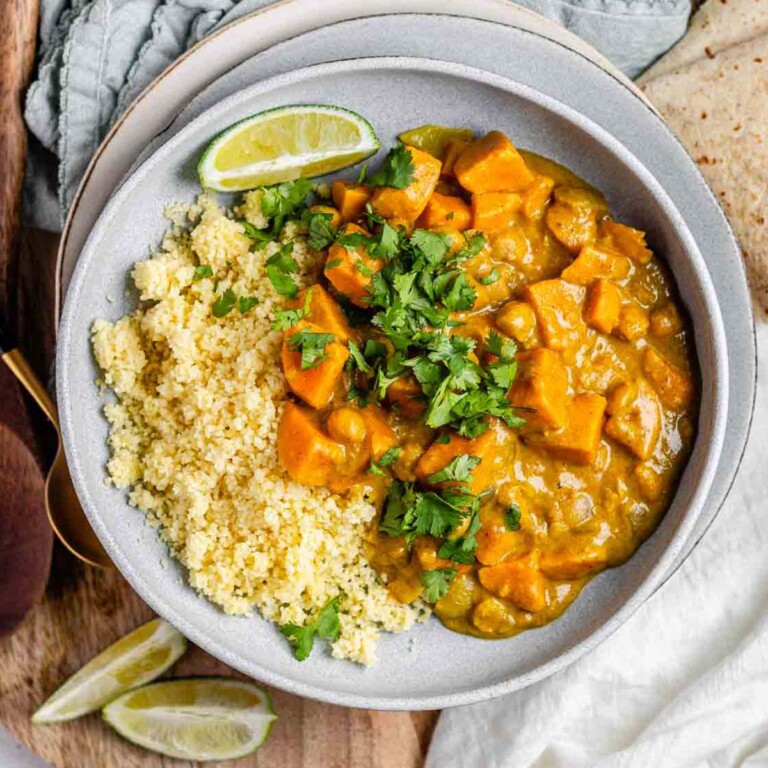 Curry with chunks of sweet potato in a bowl with couscous, a lime wedge, and cilantro.