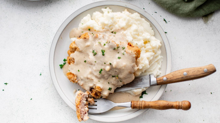 Breaded fried steak topped with gravy on a plate with mashed potatoes, a fork and knife.
