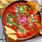 Tomato sauce with poached eggs in a skillet, topped with crumbled cheese, chopped herbs, pickled red onions, and tortilla chips.