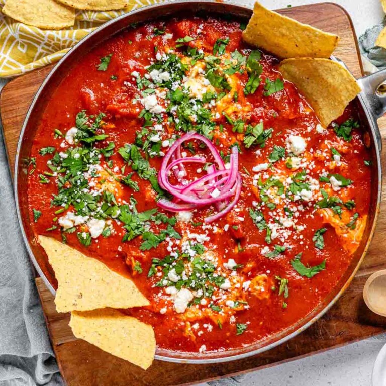 Tomato sauce with poached eggs in a skillet, topped with crumbled cheese, chopped herbs, pickled red onions, and tortilla chips.