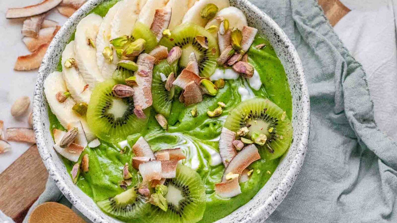 Green smoothie bowl topped with sliced banana, sliced kiwi, toasted coconut chips, and pistachios.