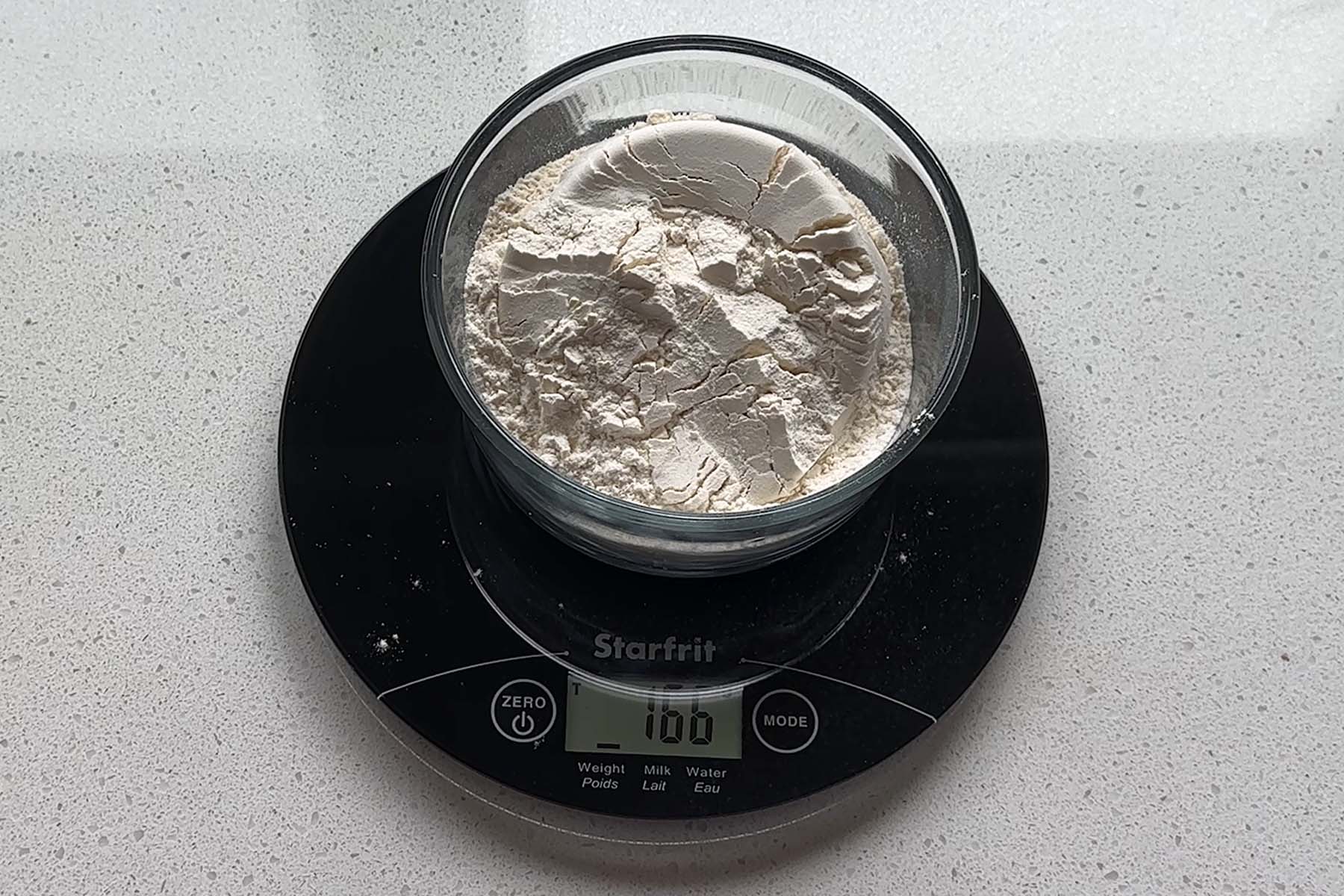Flour in a clear bowl on a kitchen scale that reads 166g.