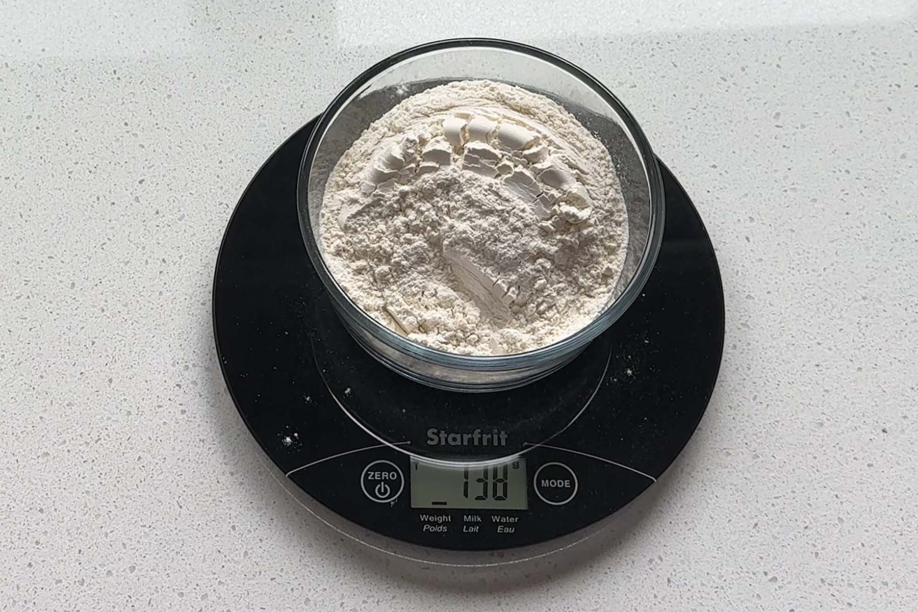 Flour in a clear bowl on a kitchen scale that reads 138g.