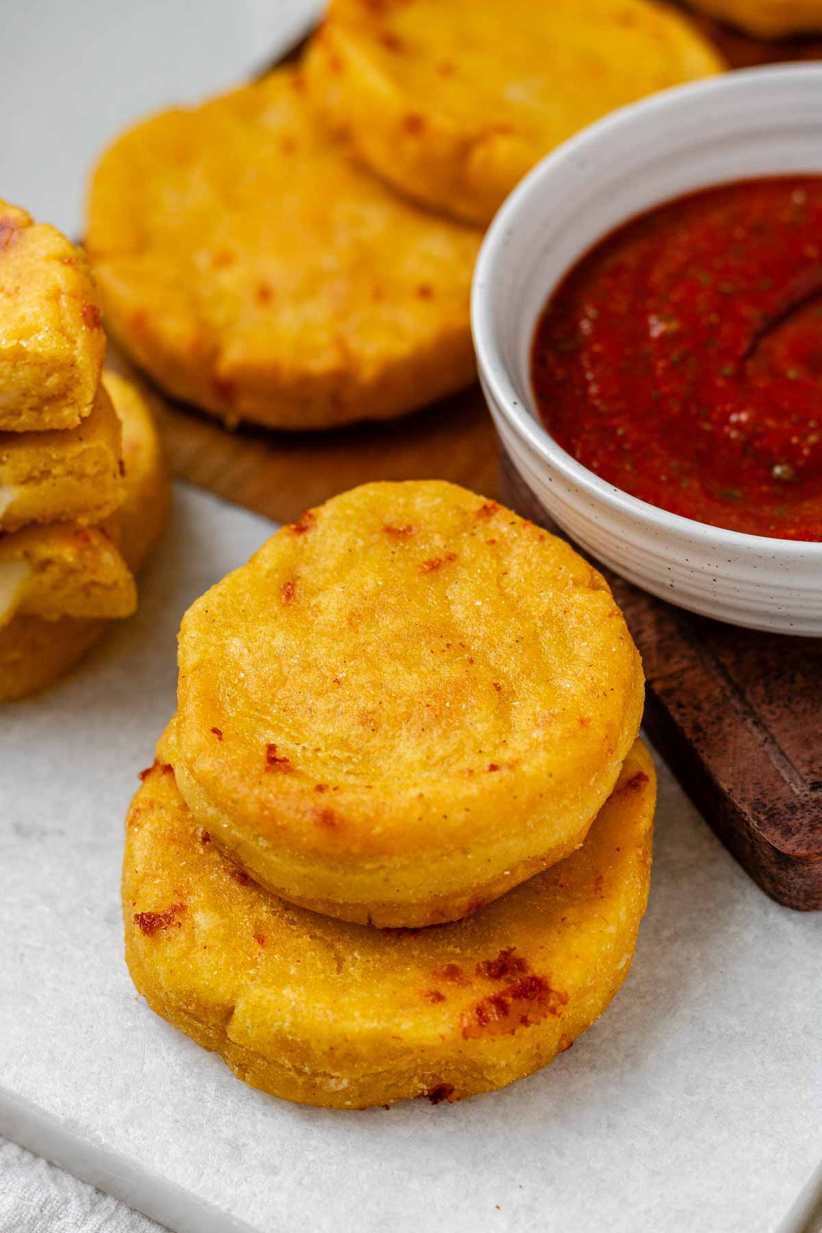 Round yellow corn dough appetizers on a platter beside a bowl of red sauce.