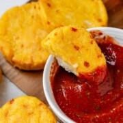 Round yellow corn dough appetizers on a platter beside a bowl of red sauce with one in the sauce bowl.