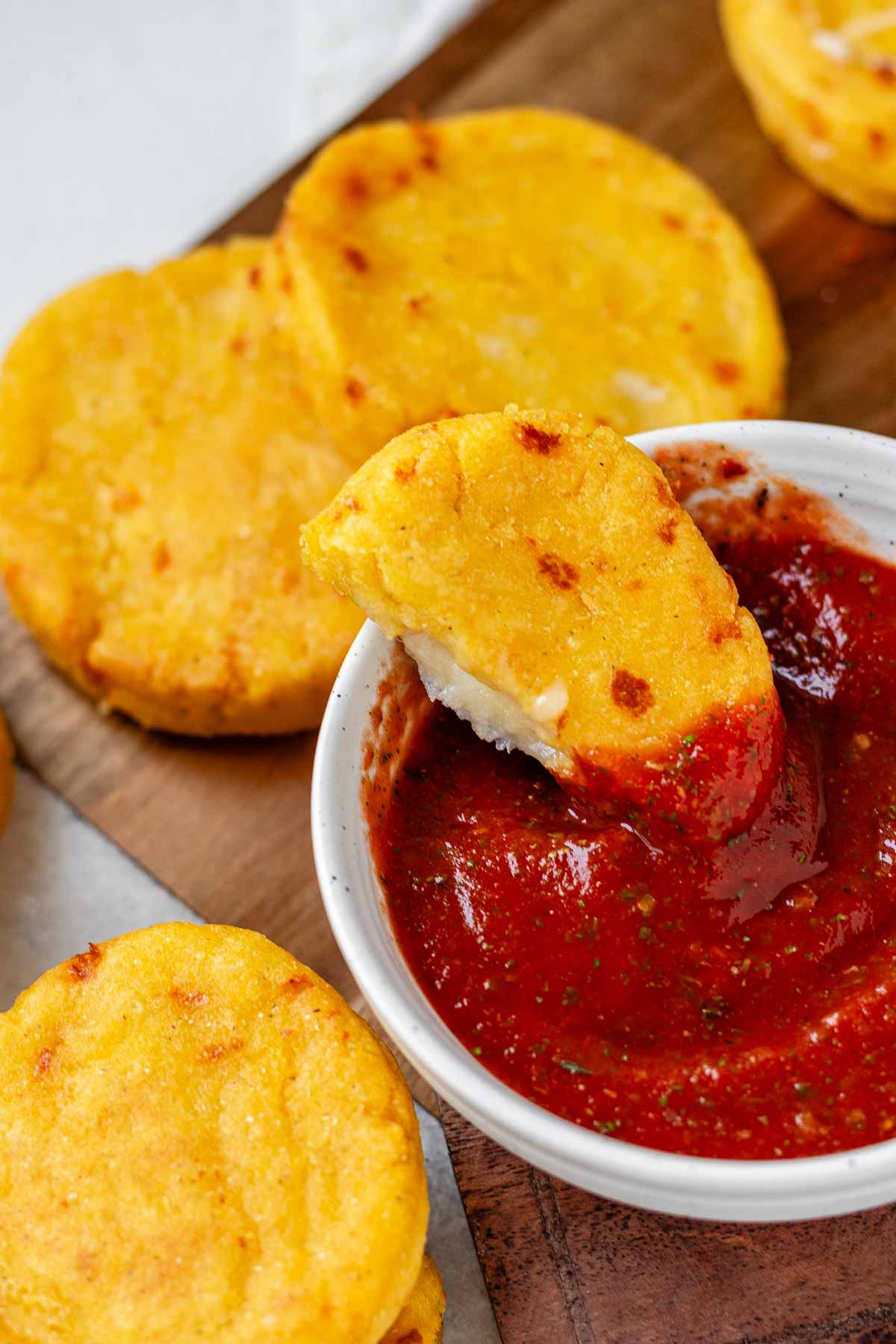 Round yellow corn dough appetizers on a platter beside a bowl of red sauce with one in the sauce bowl.