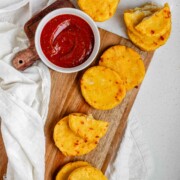 Round yellow corn dough appetizers on a platter beside a bowl of red sauce.