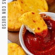 Round yellow corn dough appetizers on a platter beside a bowl of red sauce with one in the bowl and the text arepas con queso.