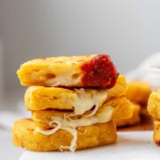 Round yellow corn dough appetizers are stacked on a platter, cut in half, one has red sauce on the end and the text arepas con queso.
