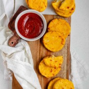 Round yellow corn dough appetizers on a platter beside a bowl of red sauce and the text arepas con queso.