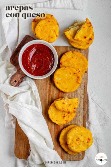 Round yellow corn dough appetizers on a platter beside a bowl of red sauce and the text arepas con queso.