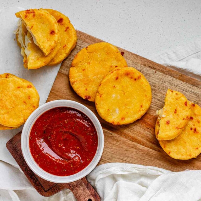 Round yellow corn dough appetizers on a platter beside a bowl of red sauce.