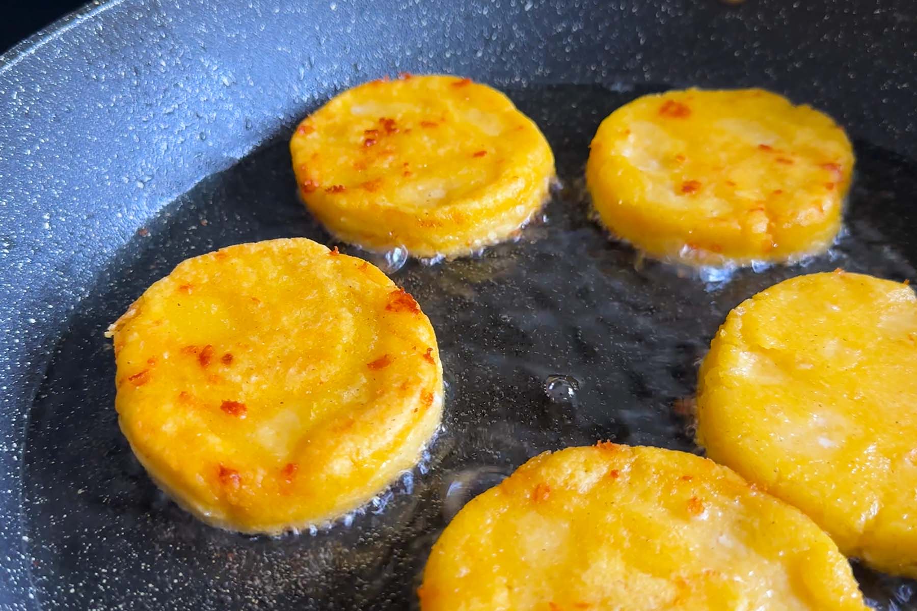 Corn dough in flattened circles in a frying pan with oil.