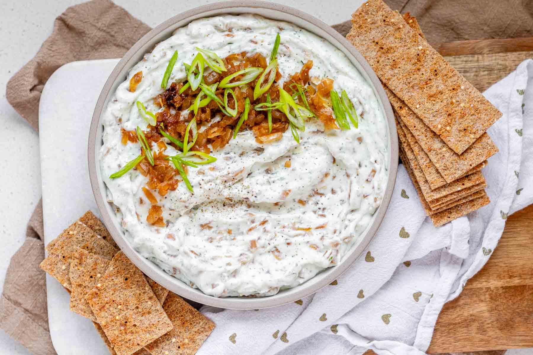 A bowl of white chunky dip with caramelized onion and sliced green onions.