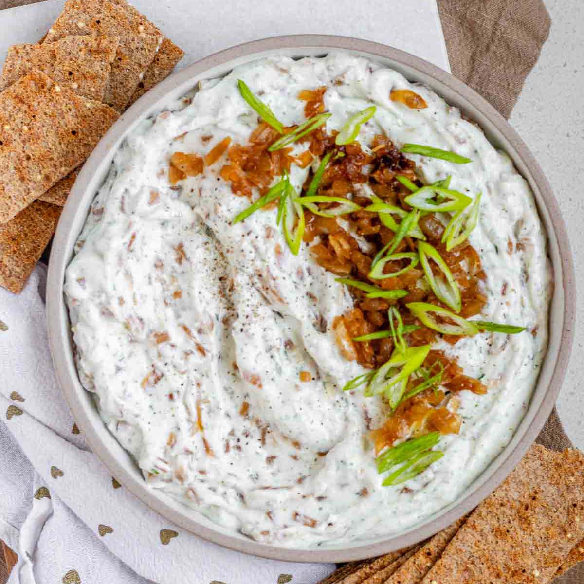 A bowl of white chunky dip with caramelized onion and sliced green onions.