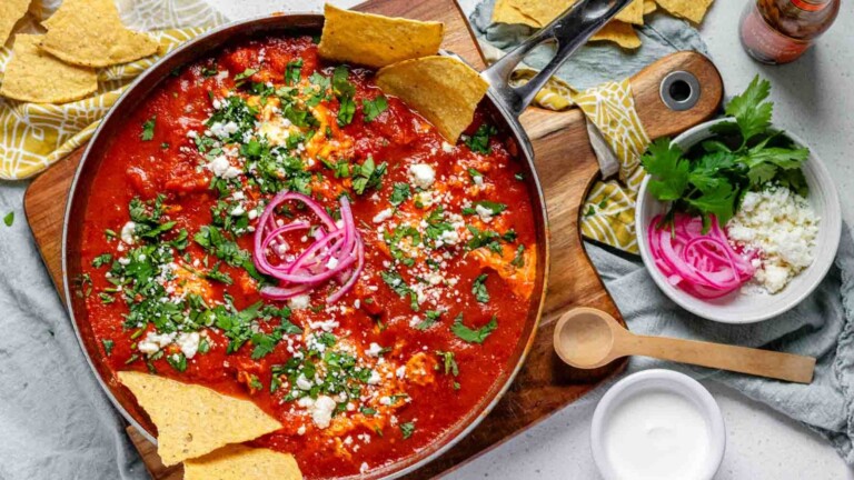 Tomato sauce with poached eggs in a skillet, topped with crumbled cheese, chopped herbs, pickled red onions, and tortilla chips.
