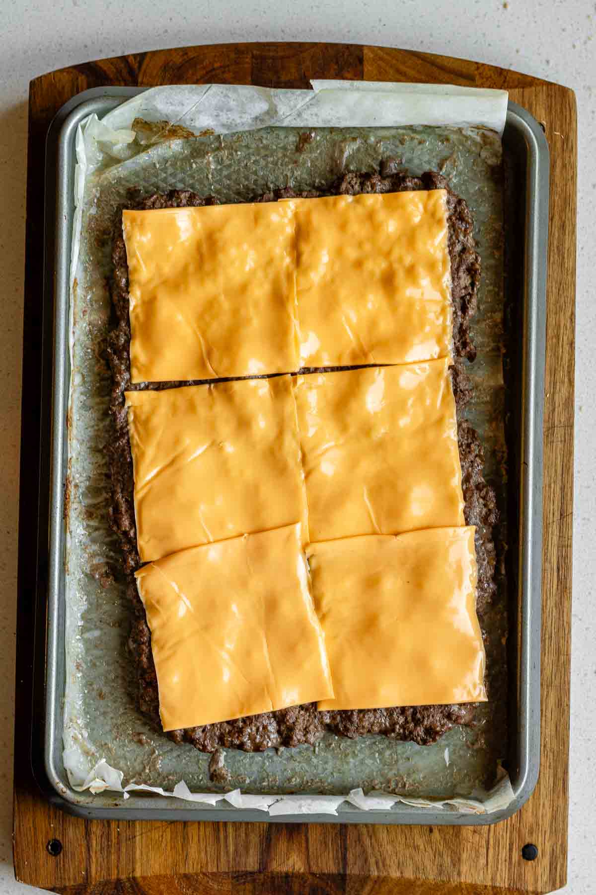 A cooked ground beef slab on a sheet pan covered in cheese slices.