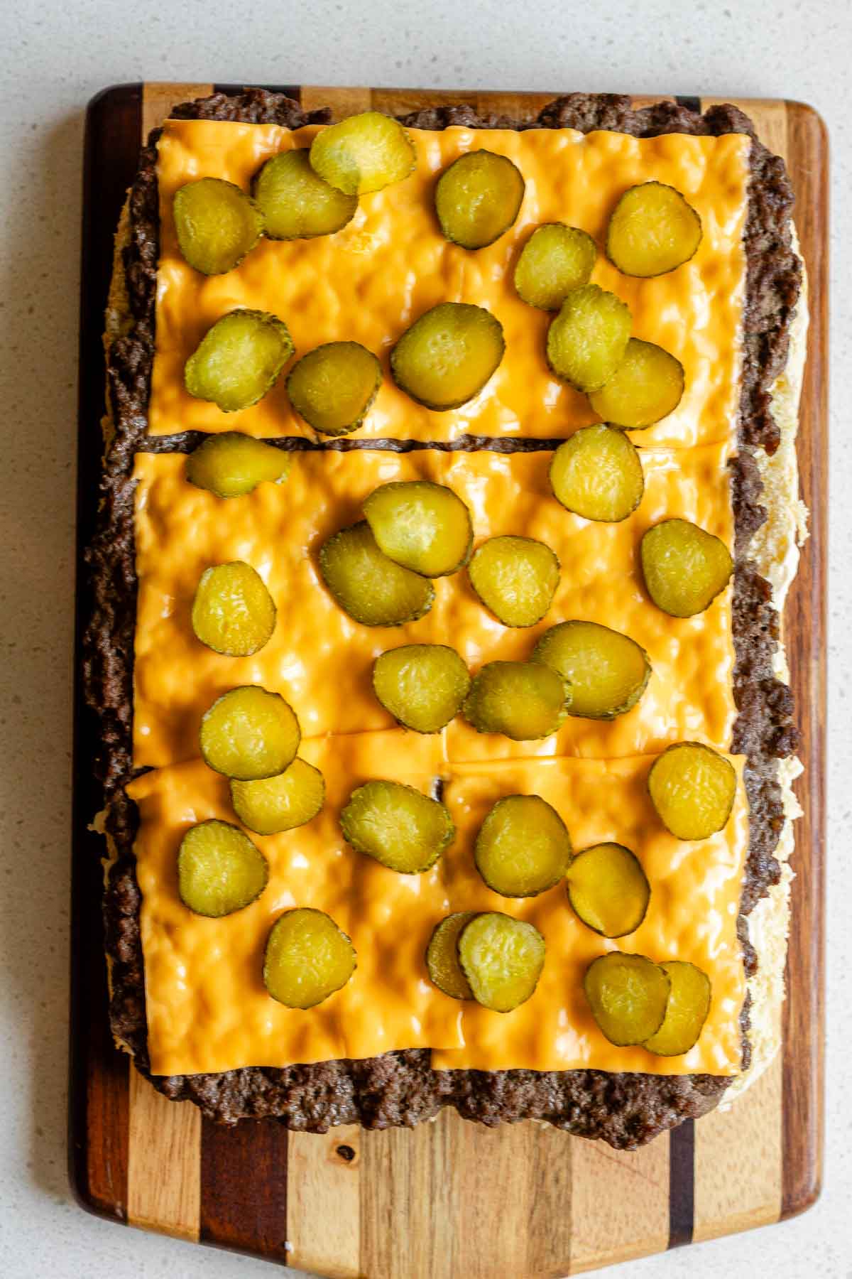 A cooked ground beef slab on a wooden board covered in cheese slices and pickles.