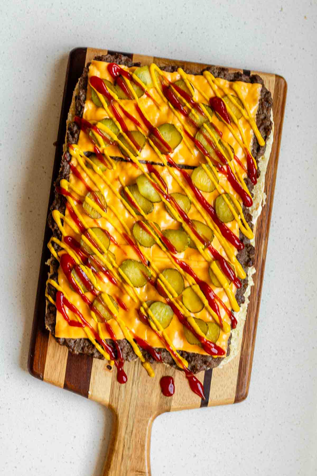 A cooked ground beef slab on a wooden board covered in cheese slices, pickles, mustard, and ketchup.