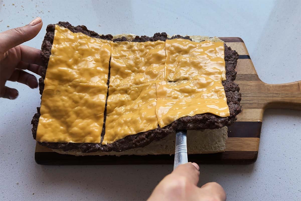 A cooked layer of beef and cheese being laid on top of buns on a cutting board.