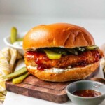 A chicken sandwich on a wooden platter beside bowls of sauce and pickles.