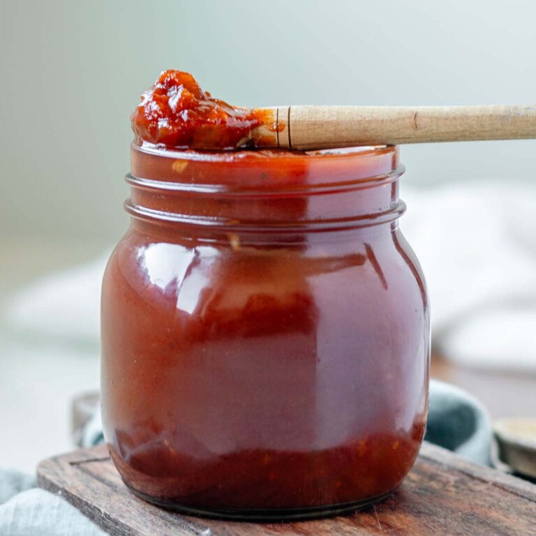 Reddish brown BBQ sauce in a jar with a spoon of sauce resting over the top.