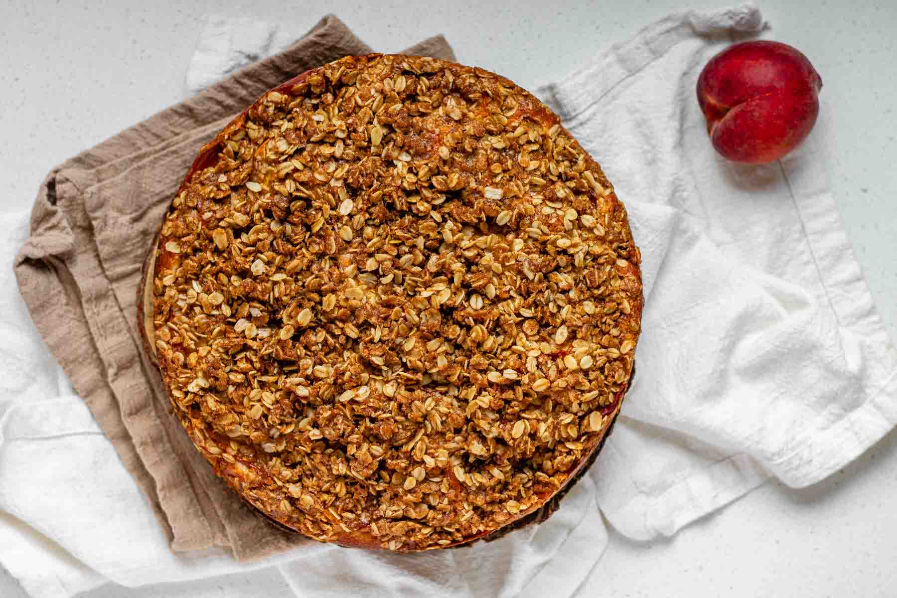 A baked cake topped with brown sugar oats beside a peach.