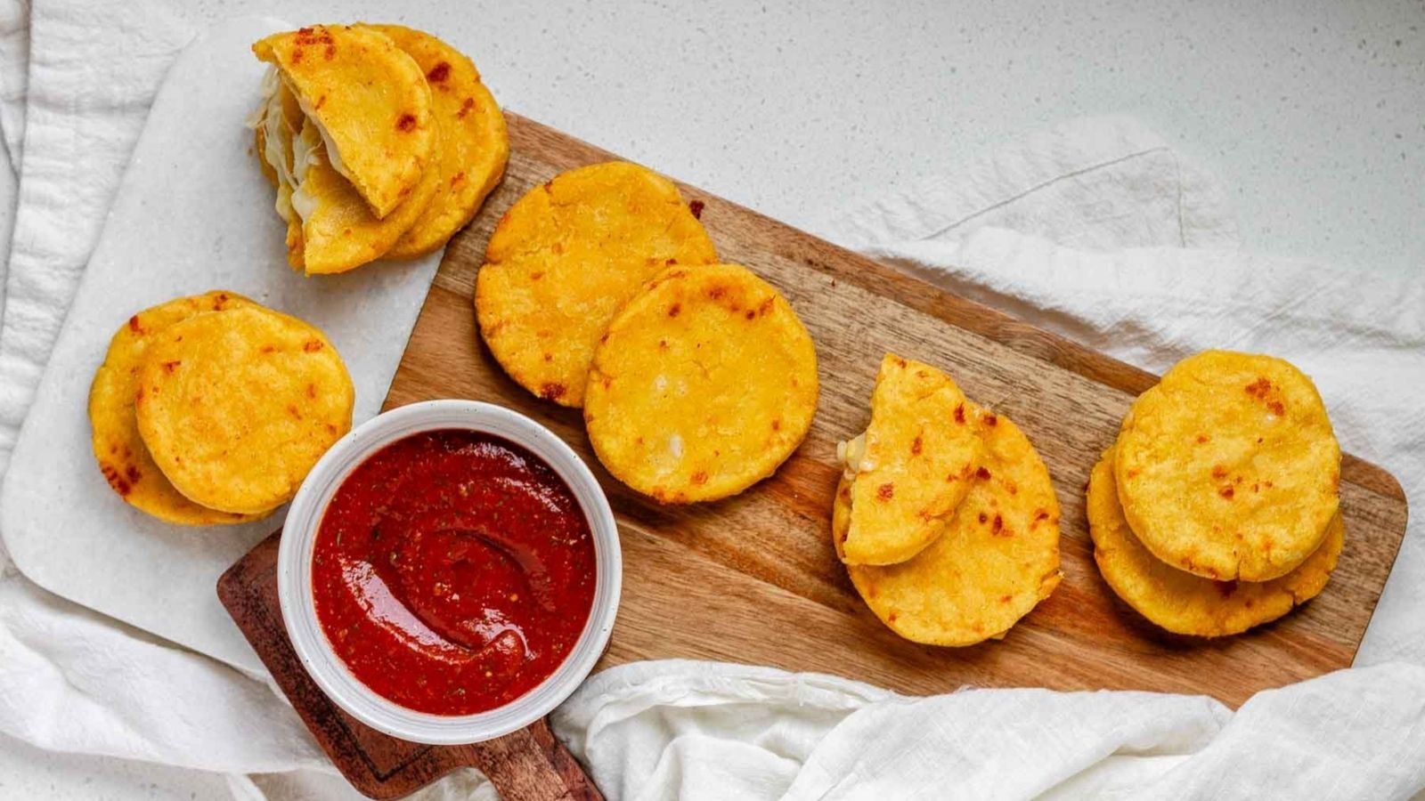 Circle corn cakes on a platter beside a bowl of red sauce.