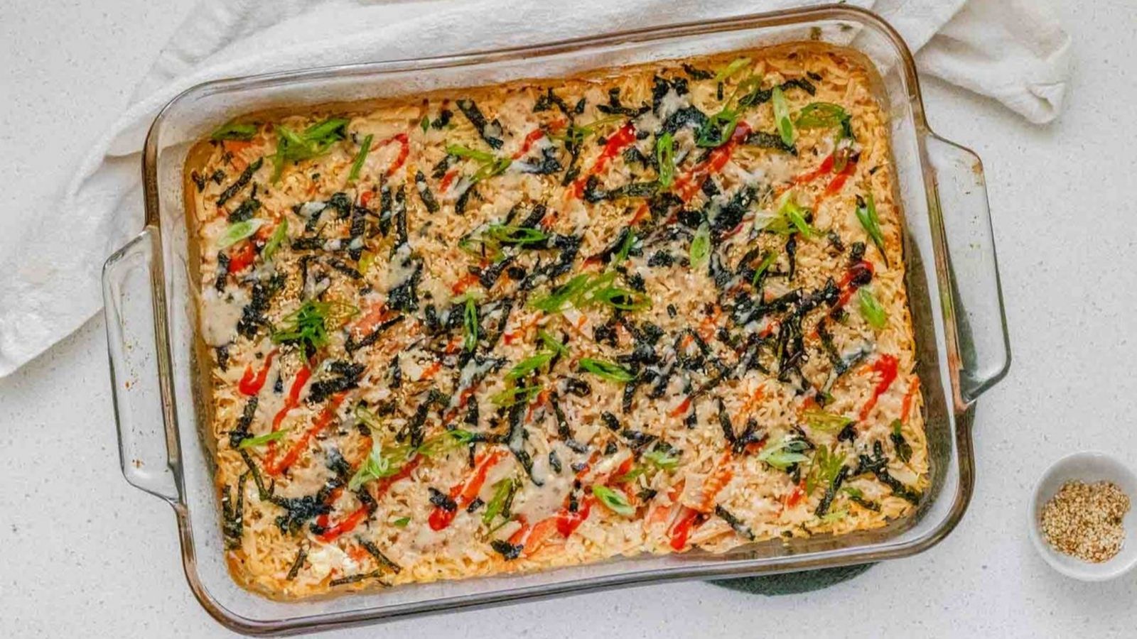 A rice casserole in a baking dish topped with nori, green onions, and a drizzle of hot sauce.