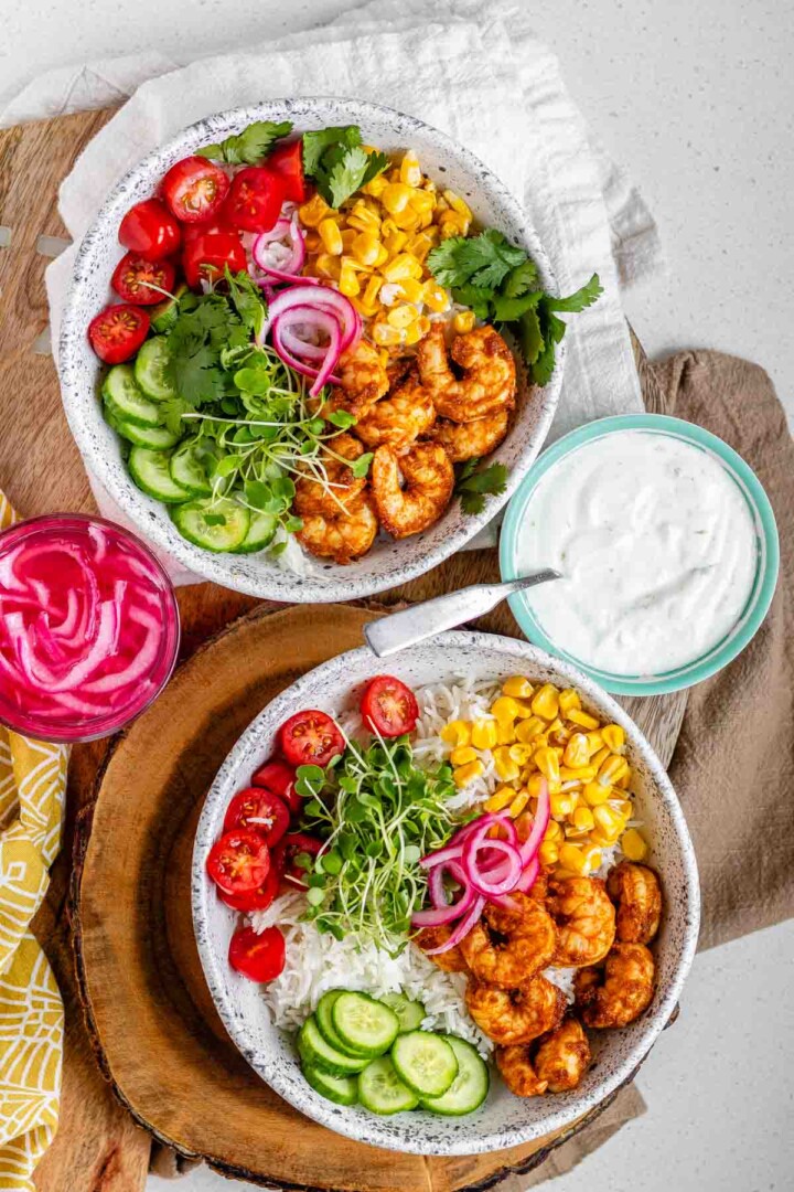 2 bowls with shrimp, cucumbers, sprouts, halved grape tomatoes, cilantro, corn, and pickled red onions.