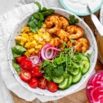 A bowl with shrimp, cucumbers, sprouts, halved grape tomatoes, cilantro, corn, and pickled red onions.