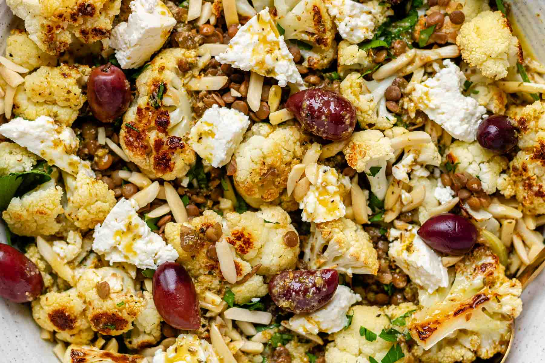 A large serving bowl with roasted cauliflower, black olives, lentils, feta cheese, slivered almonds, and fresh parsley.