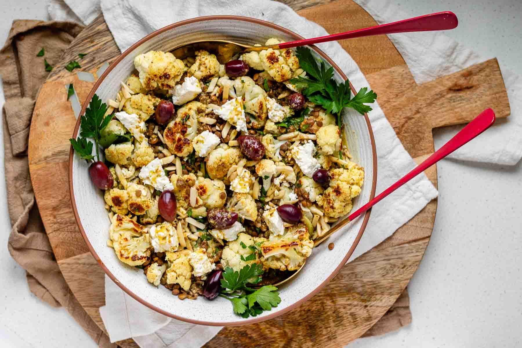 A large serving bowl with roasted cauliflower, black olives, lentils, feta cheese, slivered almonds, and fresh parsley.