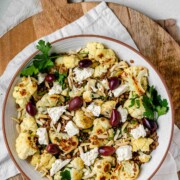 A large serving bowl with roasted cauliflower, black olives, lentils, feta cheese, slivered almonds, and fresh parsley.