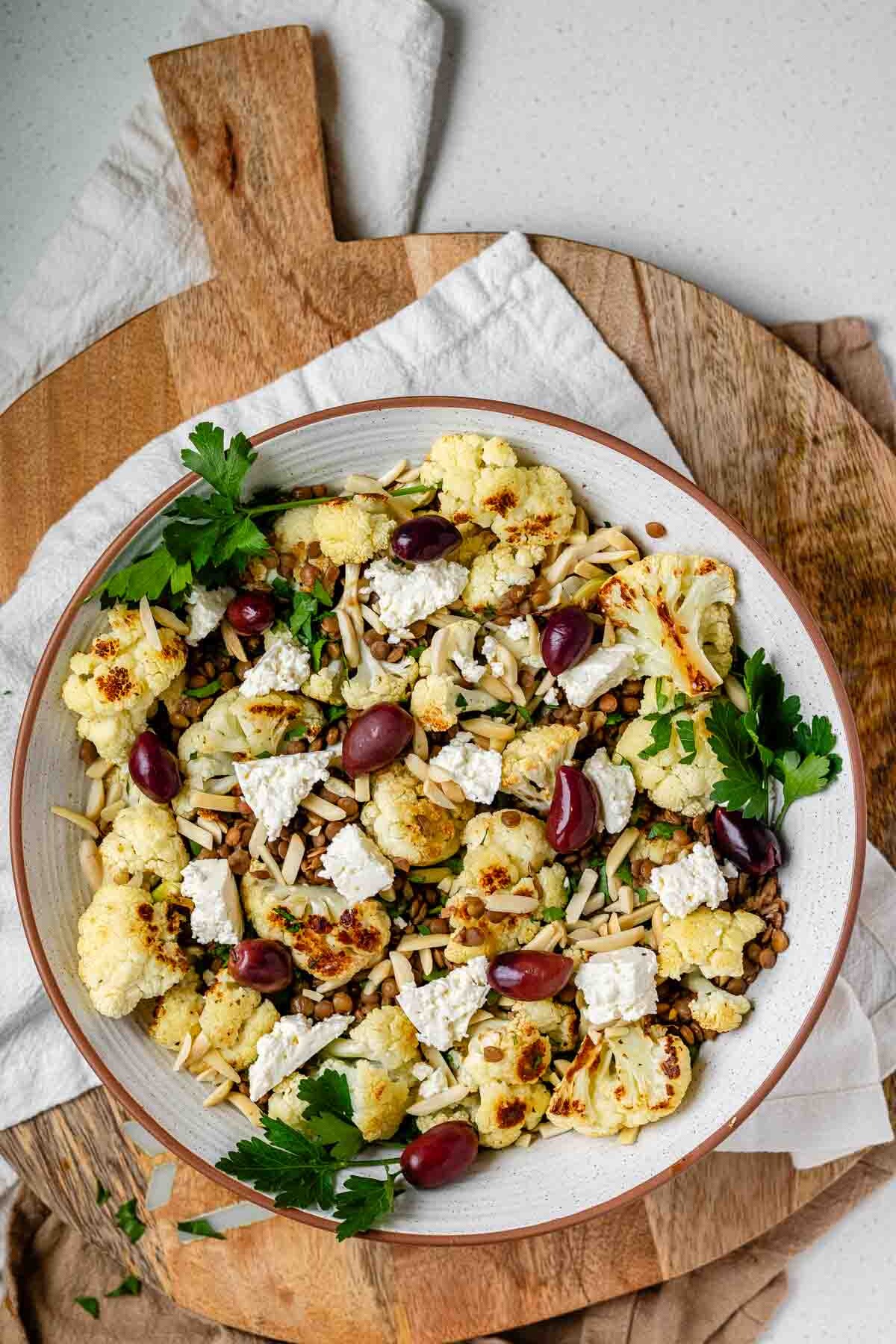 A large serving bowl with roasted cauliflower, black olives, lentils, feta cheese, slivered almonds, and fresh parsley.