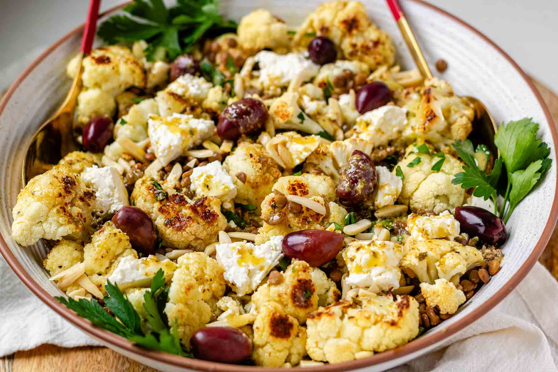 A large serving bowl with roasted cauliflower, black olives, lentils, feta cheese, slivered almonds, and fresh parsley.