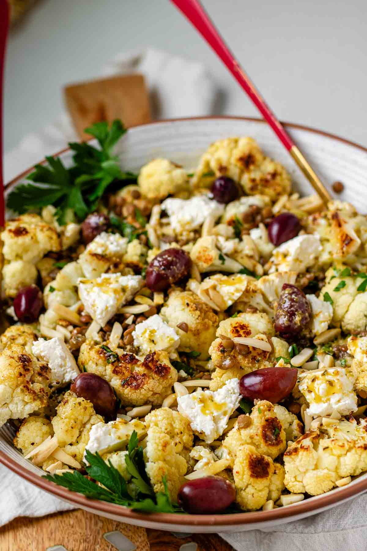 A large serving bowl with roasted cauliflower, black olives, lentils, feta cheese, slivered almonds, and fresh parsley.