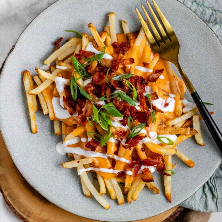 Fries on a plate topped with cheese sauce, a yogurt drizzle, crispy bacon, and sliced green onions.