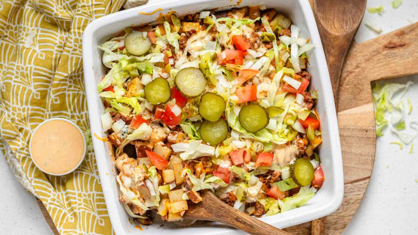 Potato cubes, ground beef, Big Mac sauce and fresh veggies in a white baking dish with a wooden spoon.