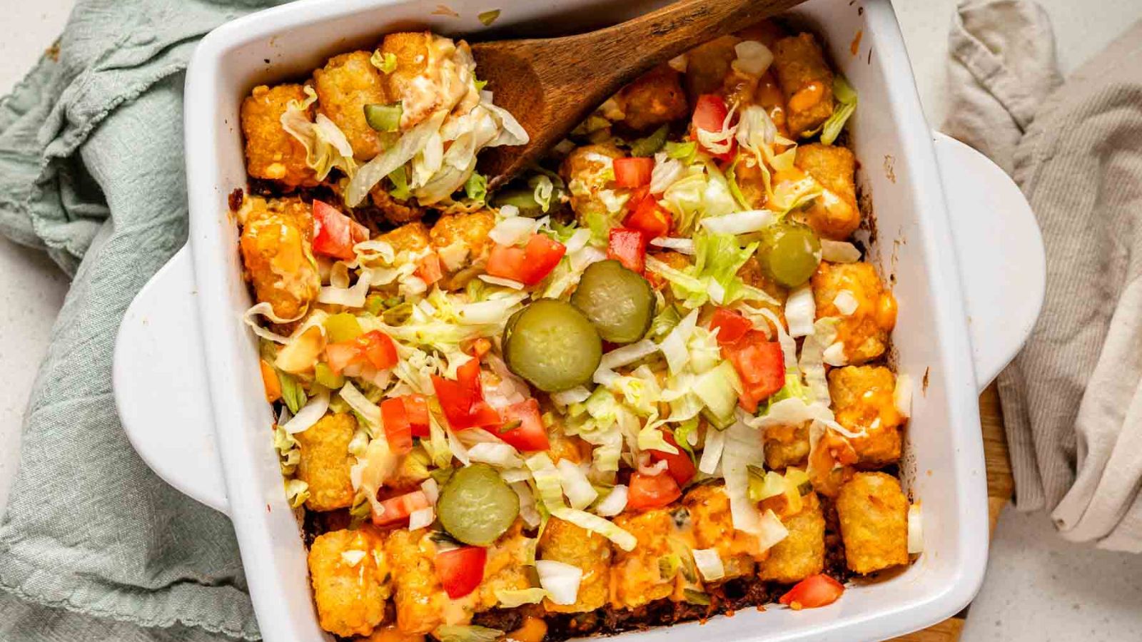 Tater tots, ground beef, Big Mac sauce and fresh veggies in a white baking dish with a wooden spoon.