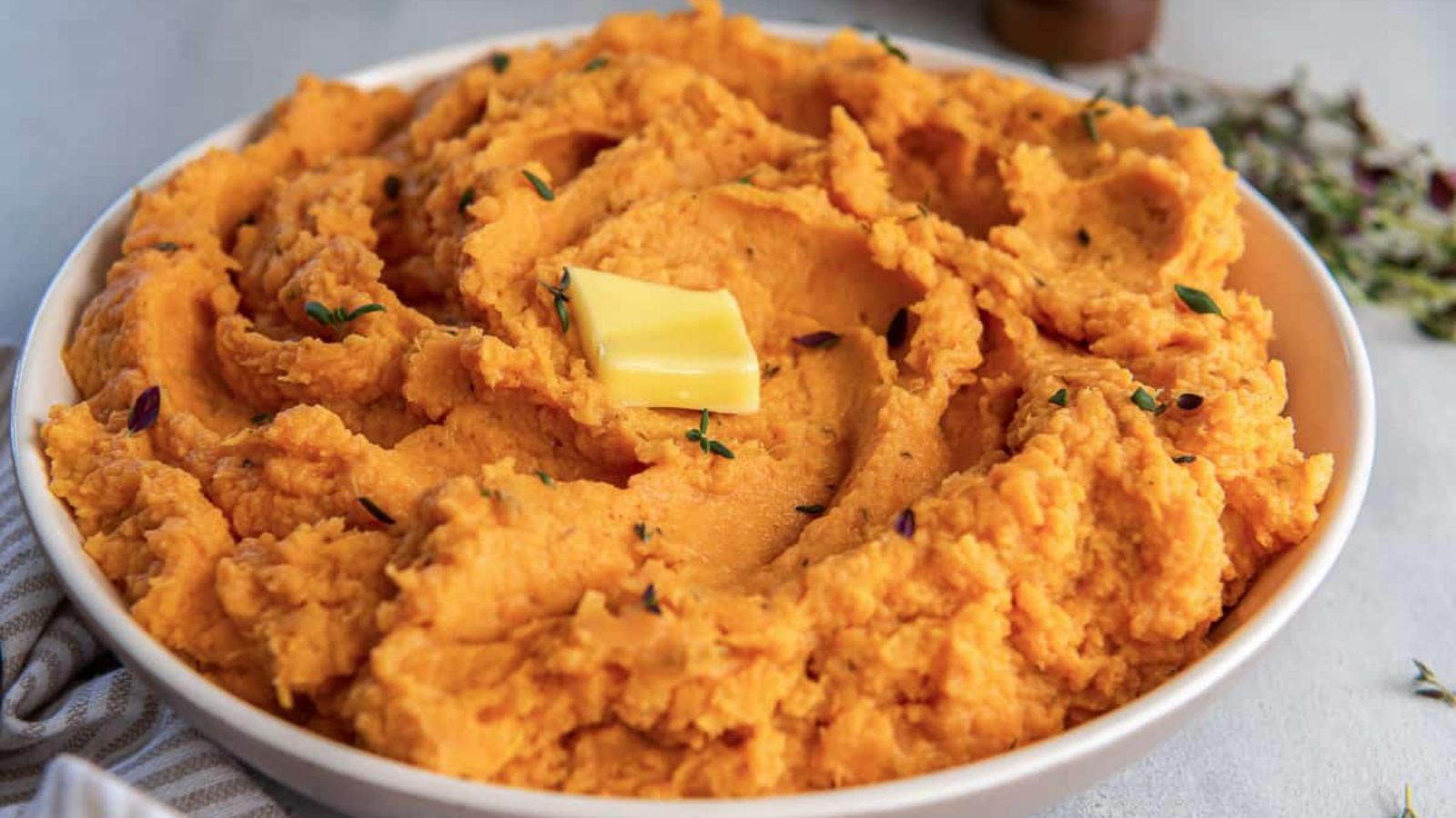 A white bowl of mashed sweet potatoes topped with butter and thyme.