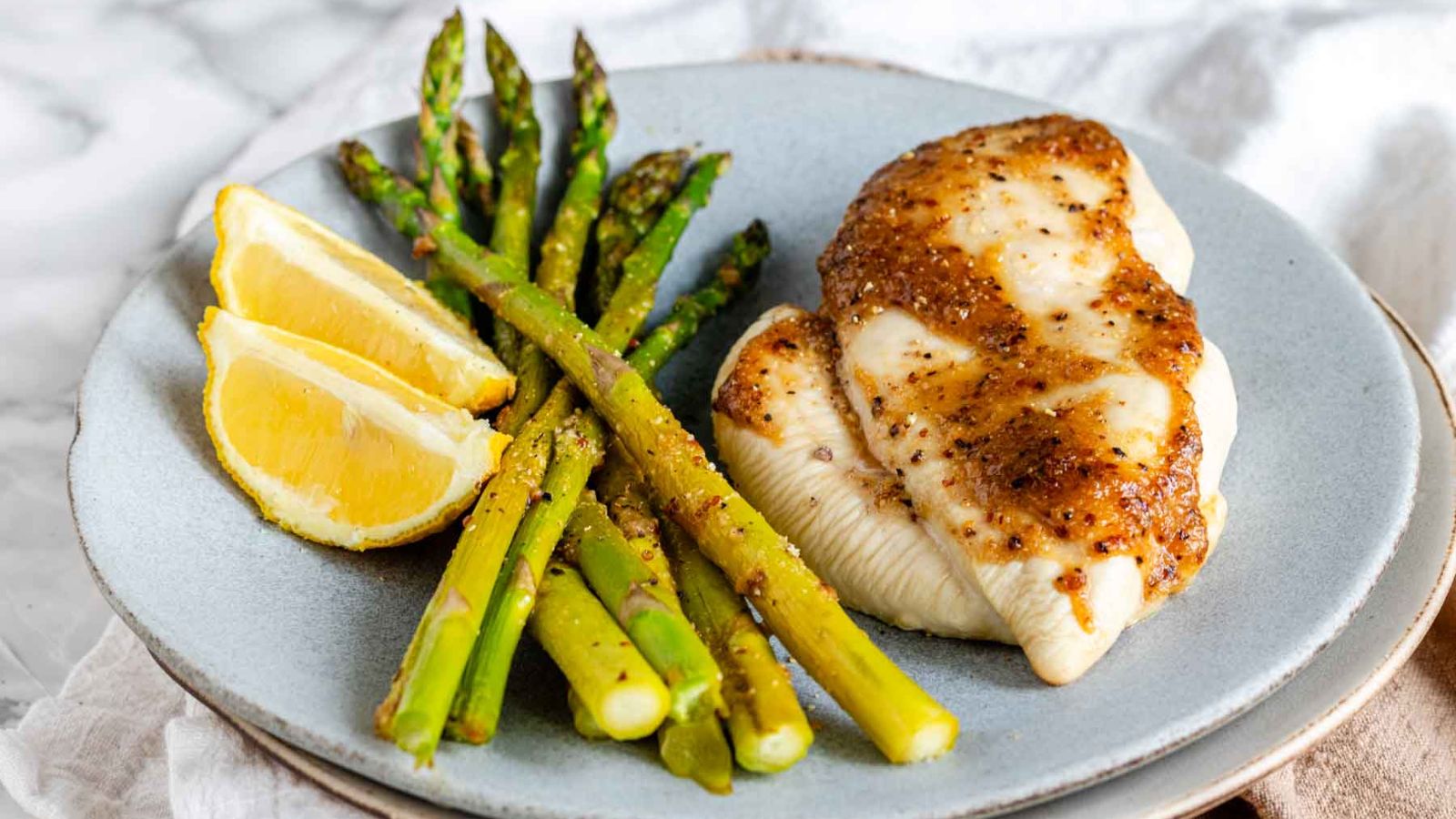 Two chicken breasts, asparagus and two lemon wedges on a light blue grey plate.